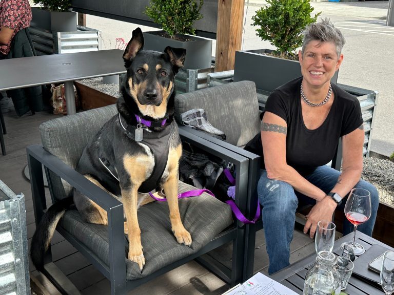 A person with short gray hair sits at an outdoor café table in Healdsburg, dressed in a black shirt and jeans. Beside them, a black and tan dog relaxes on its own chair. A wine glass sits among other items on the table. The background is filled with lush greenery, creating a serene atmosphere that feels refreshing and revitalizing.