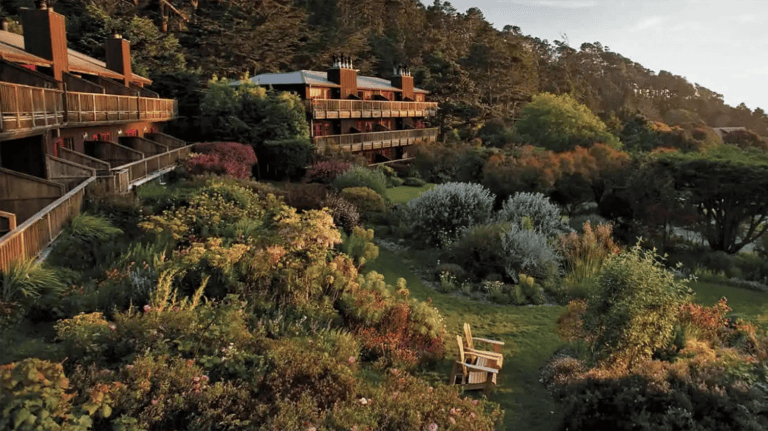 Stanford Inn offers a landscape of wooden buildings set against a rocky hillside. The area features diverse greenery, colorful flowers, and trees. A wooden bench provides a vantage point for the scene under a clear sky.