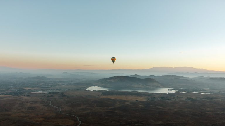 A hot air balloon drifts over a broad landscape with mountains, Lake Skinner, and wide plains during sunrise or sunset. The sky is clear, offering an unobstructed view of the peaceful scenery from above.