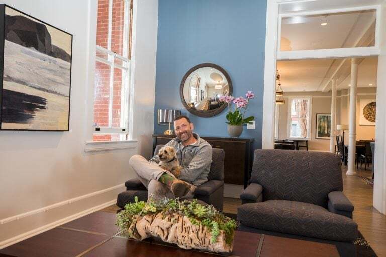 A person sits comfortably in a dimly lit chair within the warm, inviting atmosphere of the Lodge at the Presidio's living room. They hold a small dog affectionately, their smile reflecting contentment. The room is thoughtfully decorated, featuring a blue accent wall that adds depth and character. A round mirror hangs stylishly above a piece of art, adding to the aesthetic appeal. Sunlight filters through large windows, creating a natural ambiance. On the wooden table before them is an elegant arrangement of succulents, lending a fresh touch to the cozy setting.
